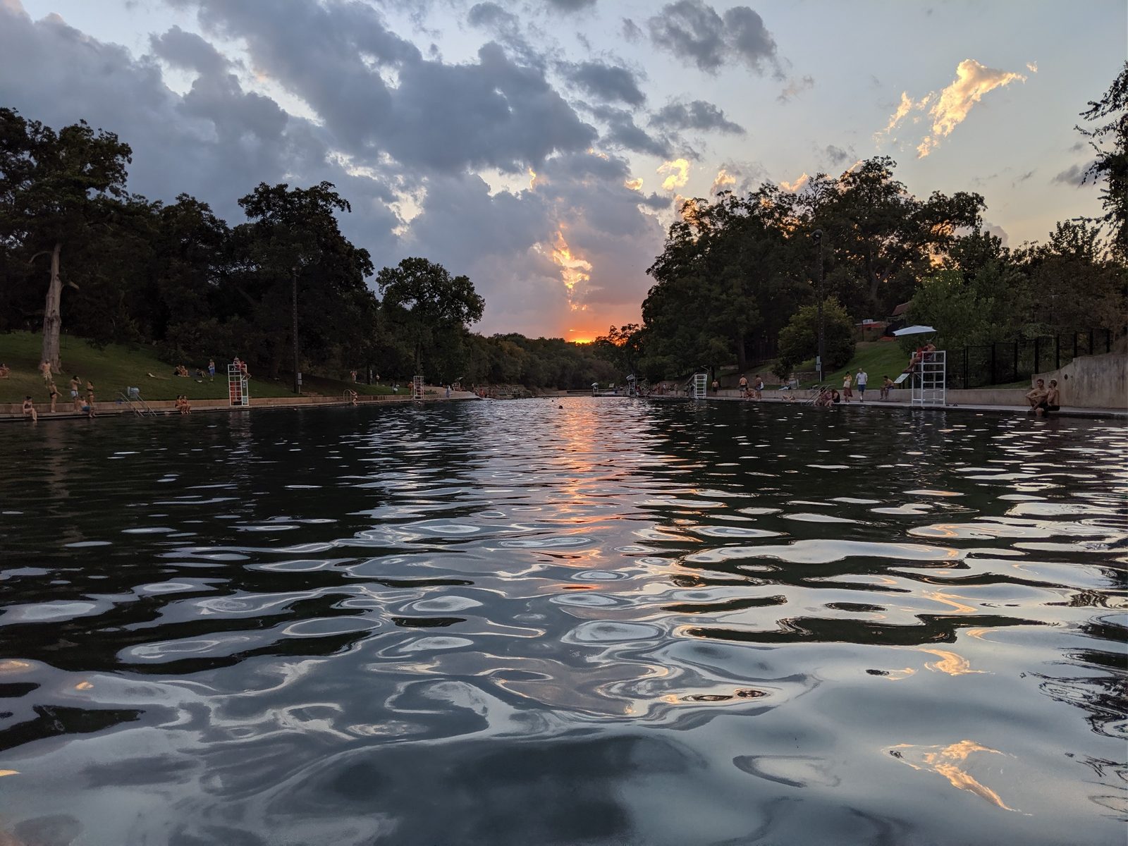 Sunset reflecting on the surface of Barton Springs Pool in Austin, Texas, with swimmers resting along the edges as storm clouds gather
